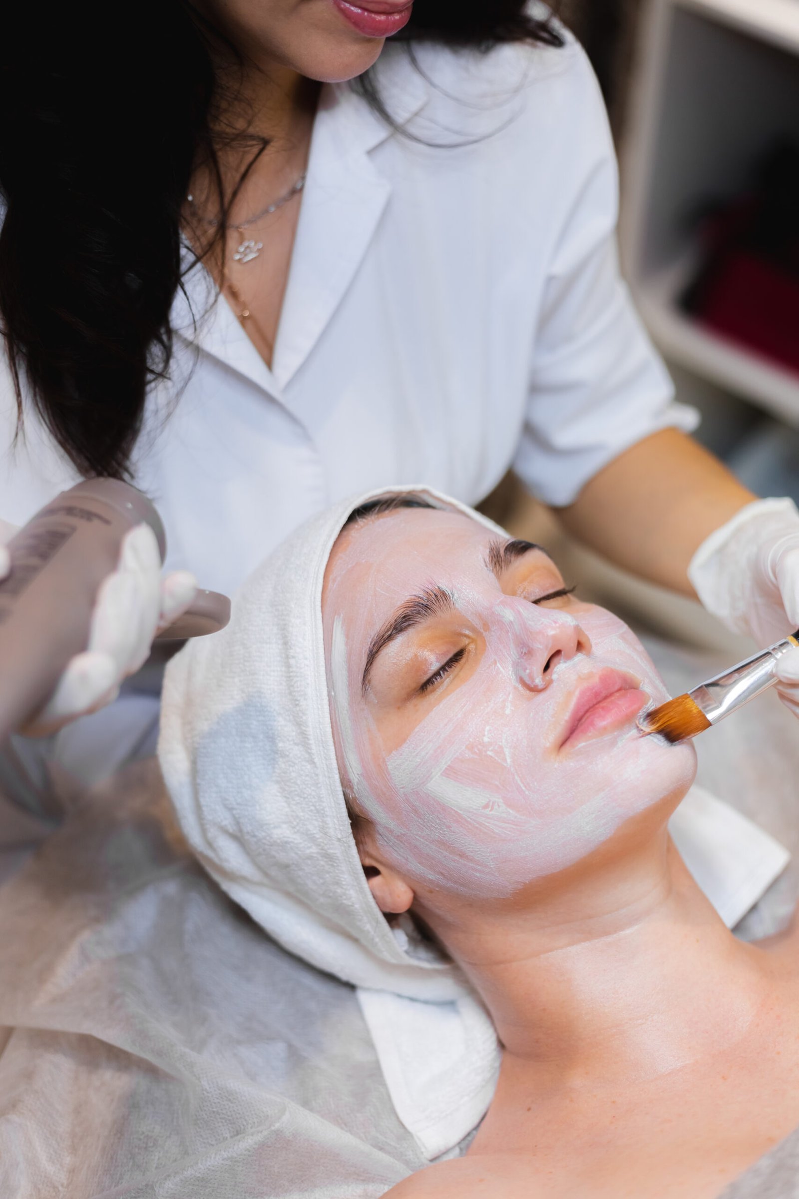 Beautician with a brush applies a white moisturizing mask to the face of a young girl client in a spa beauty salon